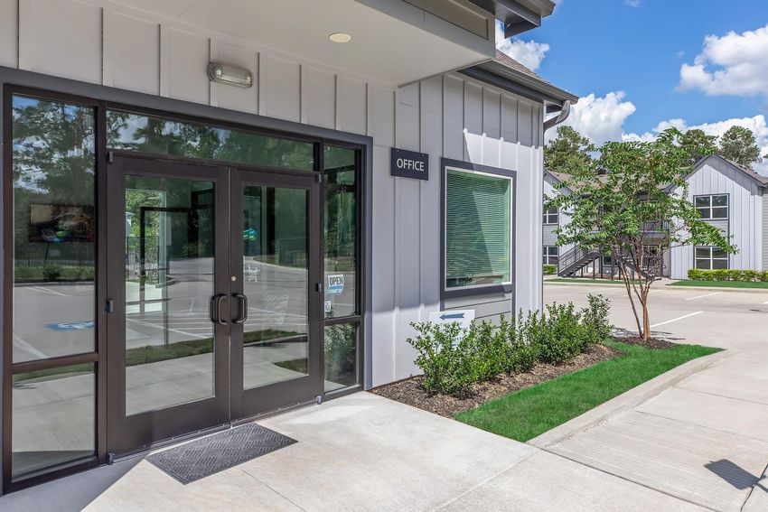Modern office entrance with double glass doors, surrounded by neatly trimmed bushes and a paved pathway. A sign indicates "OFFICE" on the wall. Bright, clear sky with a few clouds in the background, and adjacent buildings visible in the distance.