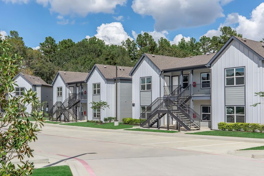 A row of modern, two-story apartment buildings with gray siding and pitched roofs, surrounded by greenery. The buildings feature balconies, large windows, and exterior staircases. The sky is bright with fluffy clouds, creating a pleasant atmosphere for the residential area.