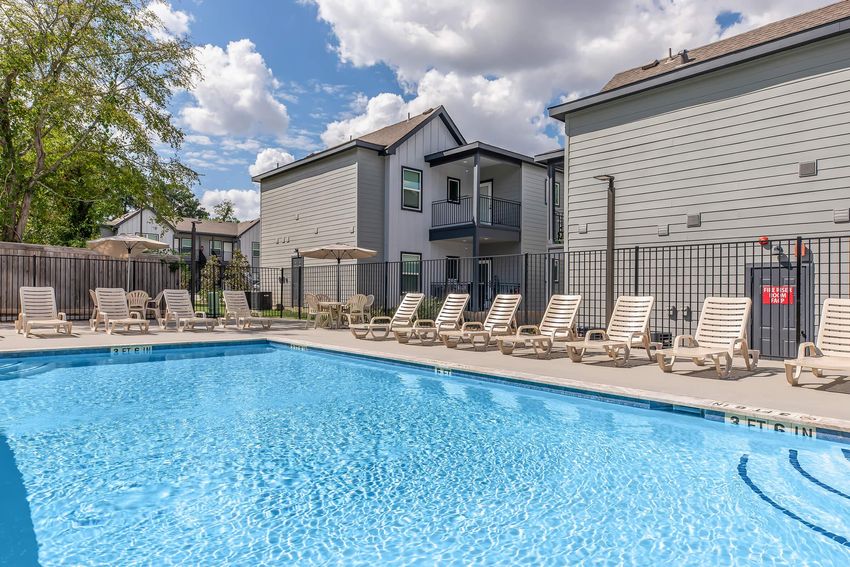 A sparkling blue swimming pool surrounded by lounging chairs, with shaded umbrellas and nearby residential buildings. Lush greenery in the background under a partly cloudy sky, creating a inviting and relaxing atmosphere for leisure and recreation.