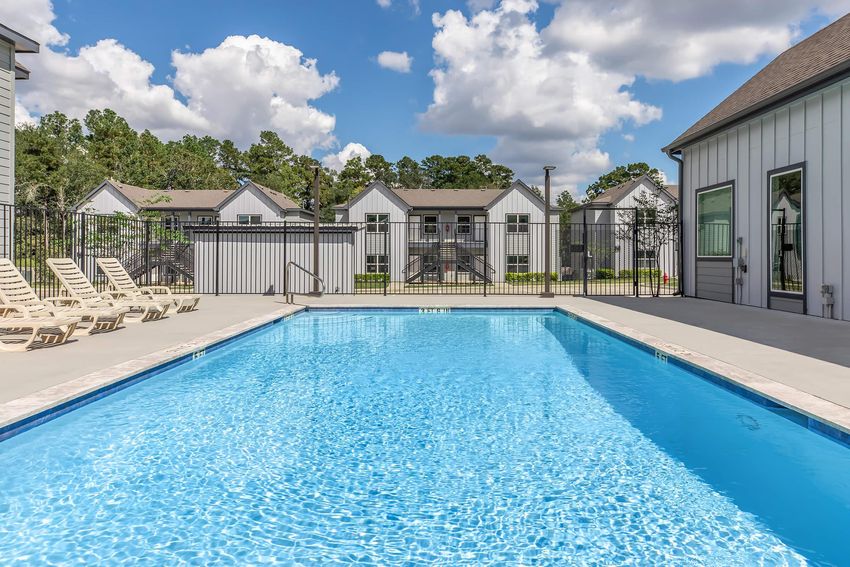 A clear swimming pool surrounded by lounge chairs, with a backdrop of modern apartment buildings and lush greenery under a partly cloudy sky. The scene conveys a relaxing outdoor atmosphere.
