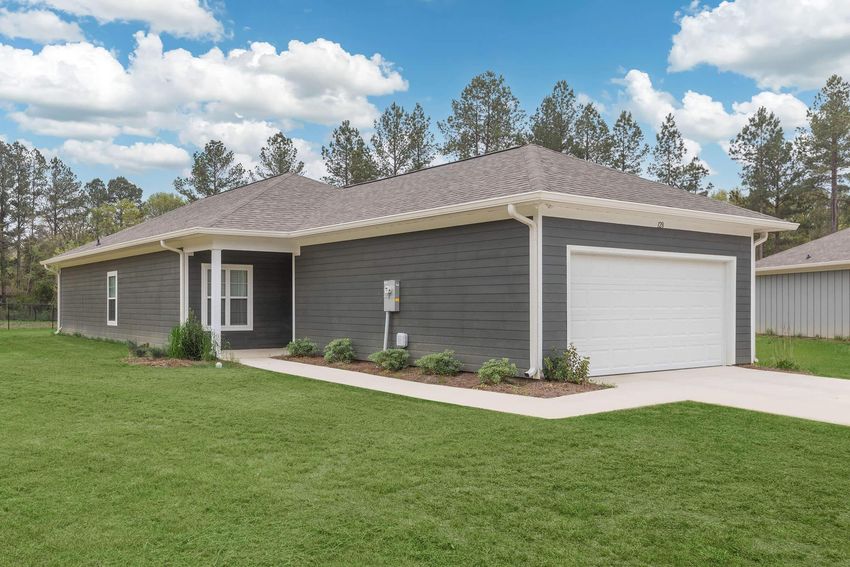 A modern, single-story house with gray siding and a white garage door, surrounded by a green lawn. The home features a covered porch area and a pathway leading to the entrance, with landscaped bushes and trees in the background under a partly cloudy sky.