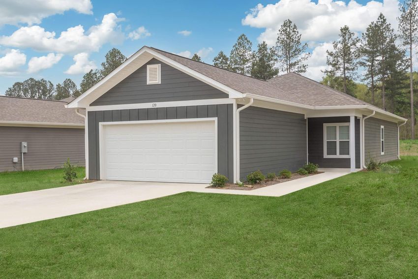 A modern single-story home with gray siding and a white garage door. The front yard is well-maintained with green grass, minimal landscaping, and trees in the background under a partly cloudy blue sky. The house features a porch and large windows.