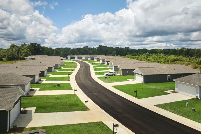 Aerial view of a suburban neighborhood featuring a curved street lined with identical single-family homes. Each house has a gray roof and a lawn, with driveways leading to garages. The scene is set under a partly cloudy sky with patches of blue. Green foliage surrounds the area, enhancing the residential atmosphere.