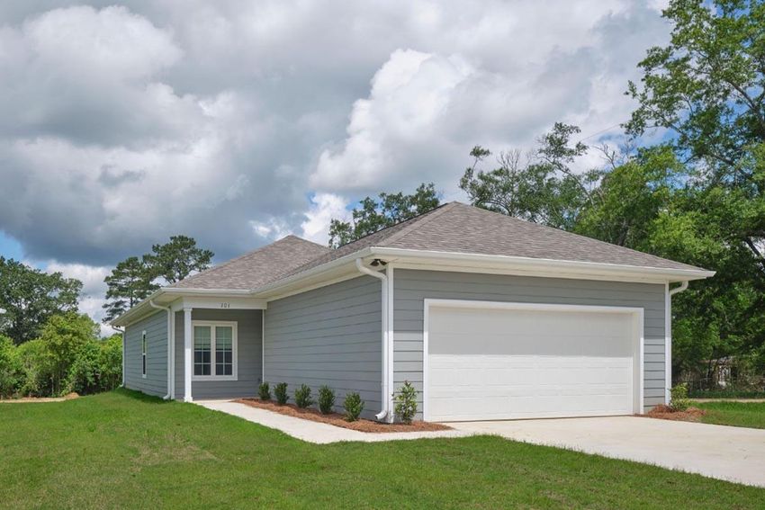 Single-story gray house with a neatly landscaped front yard, featuring a driveway leading to a garage. The home has a peaked roof, large windows, and is surrounded by trees under a partly cloudy sky. The overall setting conveys a suburban residential environment.