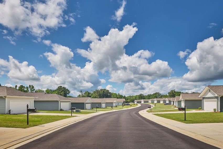 A peaceful suburban street curves through a residential neighborhood, lined with identical single-story houses under a bright blue sky filled with fluffy white clouds. The asphalt road is freshly paved, with mailboxes positioned along the neatly landscaped yards. The scene conveys a sense of tranquility and community.