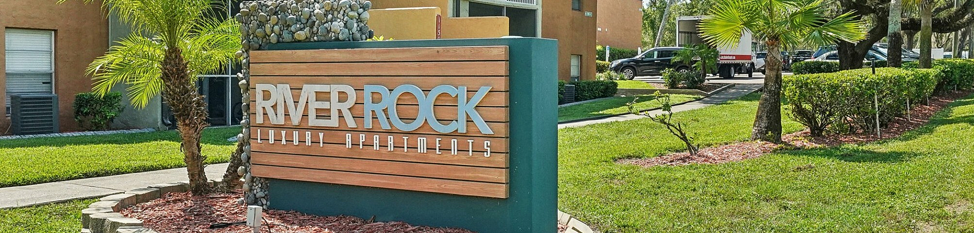Charming exterior view of River Rock Luxury Apartments with lush greenery and attractive landscaping, showcasing a welcoming entrance sign.