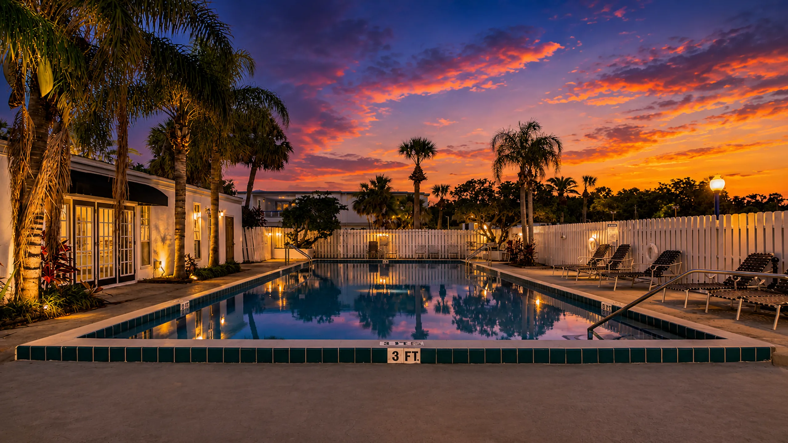 Charming poolside view at sunset, featuring a beautifully lit swimming pool surrounded by lush palm trees and cozy lounge chairs, perfect for relaxing evenings.