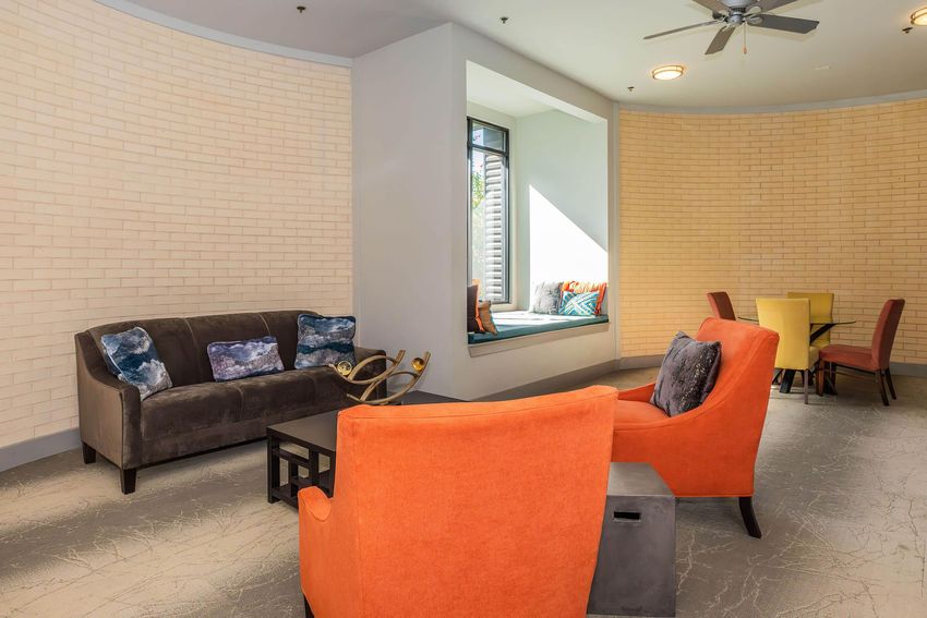 A cozy lounge area featuring a gray sofa with decorative pillows, two bright orange chairs, and a dark coffee table. In the background, a window nook with cushions invites relaxation, alongside a dining table with colorful chairs. The space is well-lit with light beige brick walls and a ceiling fan.