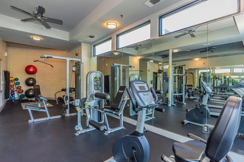 A spacious gym area featuring various fitness equipment, including weight machines, a recumbent bike, and free weights. The room is well-lit with large windows and mirrors lining one wall, creating an open and inviting environment for exercise.