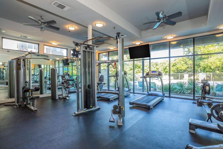 A well-lit, modern gym featuring a variety of fitness equipment, including a treadmill, weight machines, and free weights. Large windows allow natural light to fill the space, with a view of greenery outside. The room has a clean, contemporary design with ceiling fans for ventilation.