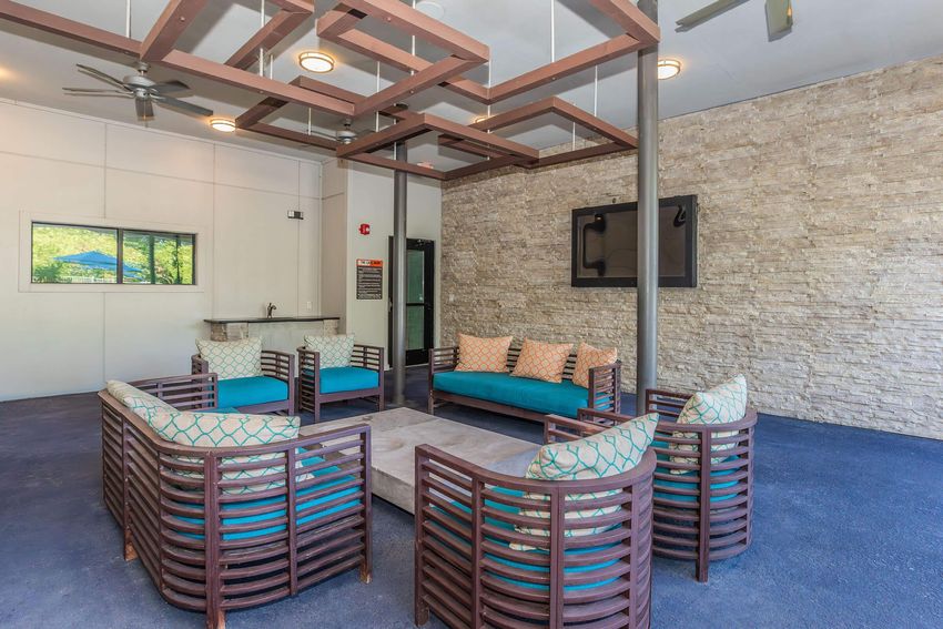 A modern lounge area featuring a circular arrangement of comfortable seating with turquoise cushions and decorative pillows. The space includes a stone wall, a ceiling with stylish light fixtures, and a bar counter in the background, offering a cozy and inviting atmosphere.
