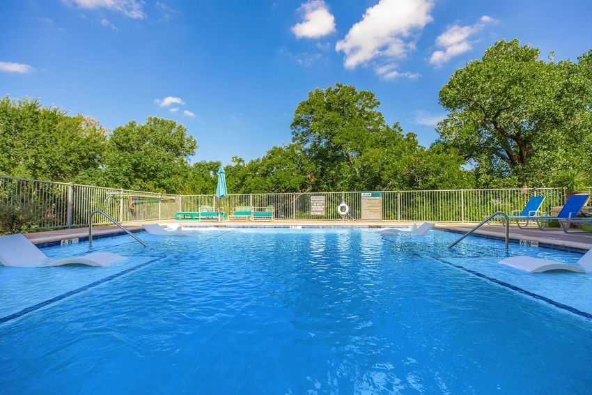 A clear blue swimming pool surrounded by green trees and a fence. The pool features a set of steps leading into the water and lounge chairs arranged nearby. A sun umbrella is visible in the background under a sunny sky with a few clouds.