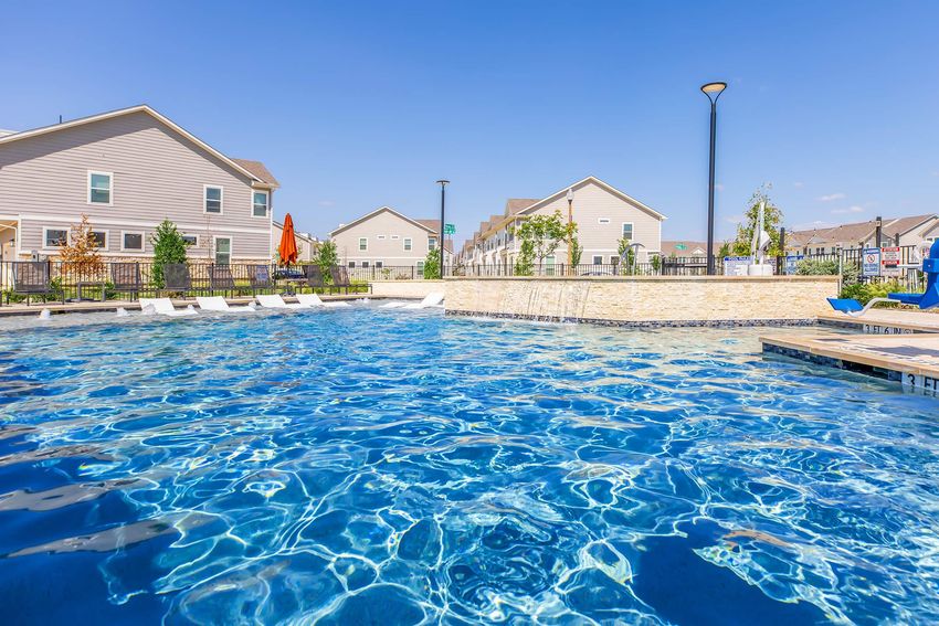 A clear blue pool with shimmering water, surrounded by lounge chairs and greenery. In the background, there are modern residential buildings and a lamp post under a bright blue sky with a few clouds. The scene is inviting and ideal for relaxation.