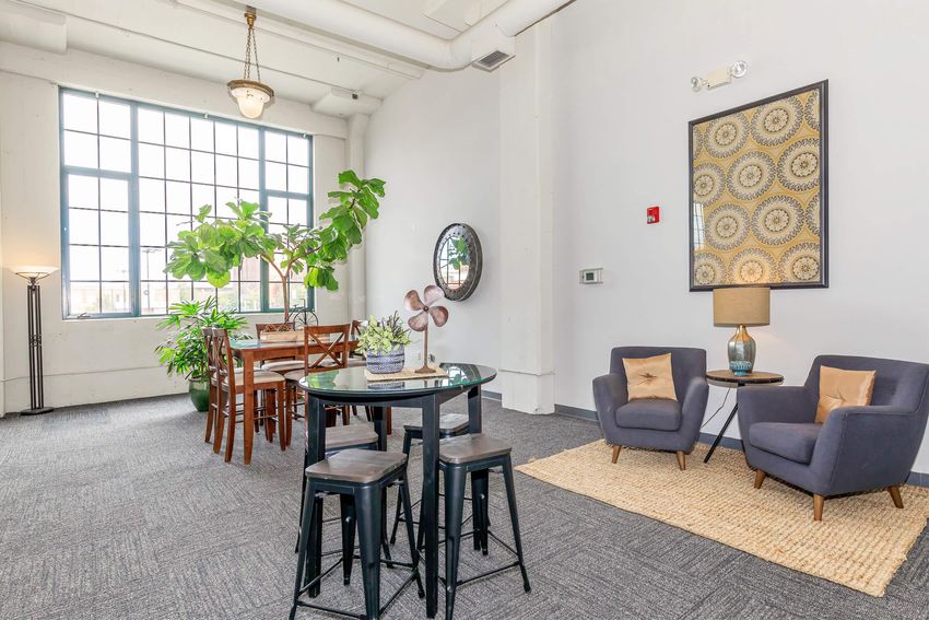 A bright, airy interior featuring a dining area with a large window, a round table surrounded by black stools, and a cozy seating area with two armchairs. A decorative rug lies on the floor, and a potted plant adds greenery, while wall art and a mirror enhance the decor. Soft lighting creates a welcoming atmosphere.