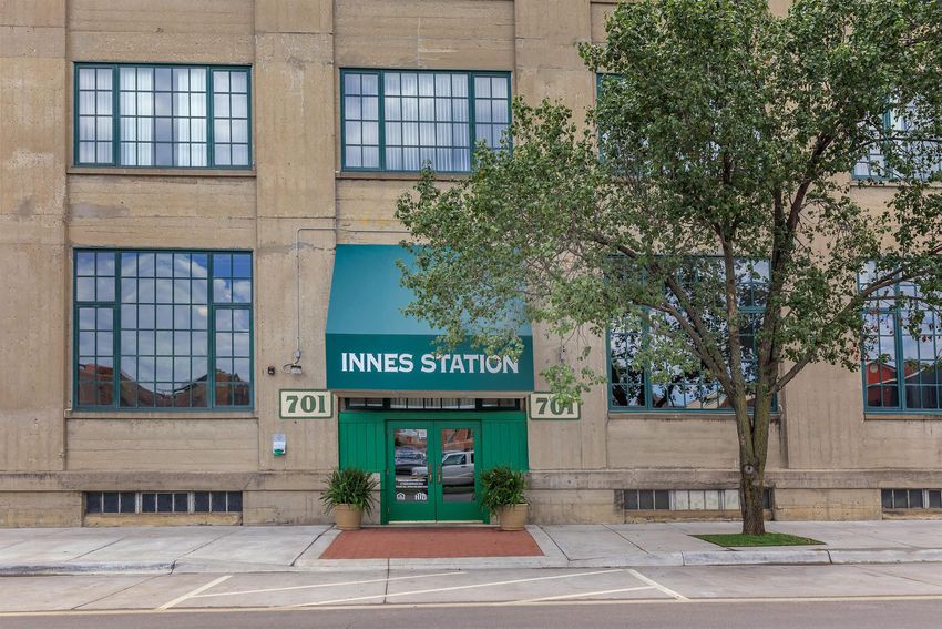 A view of Innes Station, a building featuring large windows and a green awning. The entrance is marked by the name "INNES STATION" and the address "701." Flanking the entrance are potted plants, and the facade displays a vintage industrial style with concrete walls. The surrounding area includes a sidewalk and a glimpse of the street.