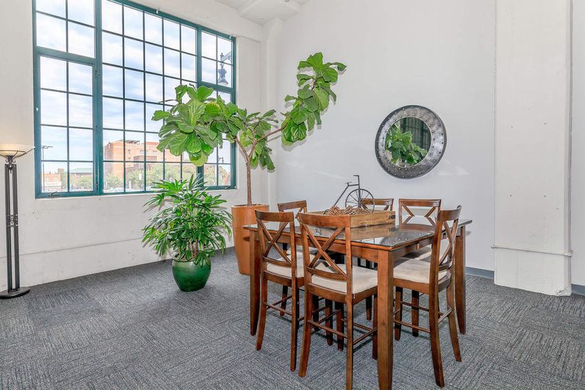 A modern dining area with a wooden table surrounded by tall chairs. A large green plant and a smaller potted plant are in the corners. A round mirror hangs on the wall, reflecting a view of the city outside through a large window with multiple panes. The floor is covered with gray carpet.