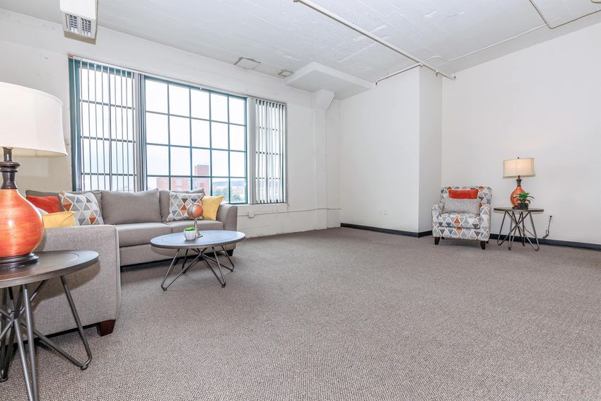 A modern, well-lit living room featuring a gray sofa with colorful cushions, an accented chair with a geometric pattern, and a coffee table. The room has large windows with vertical blinds, beige carpet, and minimalistic decor, creating a spacious and inviting atmosphere.