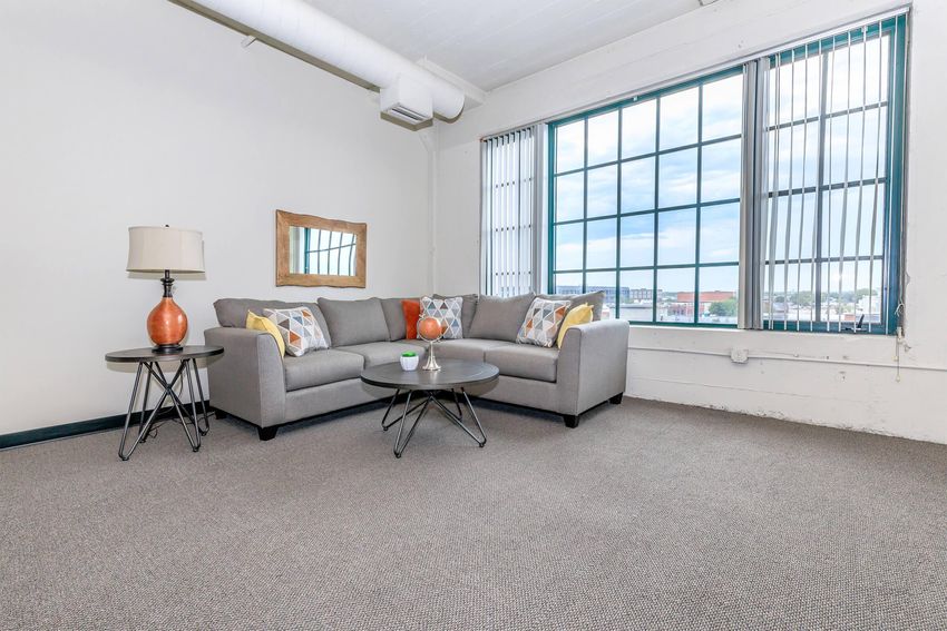 A spacious living room featuring a gray sofa with colorful accent pillows, a round coffee table, and a lamp. Large windows provide natural light, revealing a view of the cityscape outside. The room has a minimalist design with neutral colors and a clean carpeted floor.
