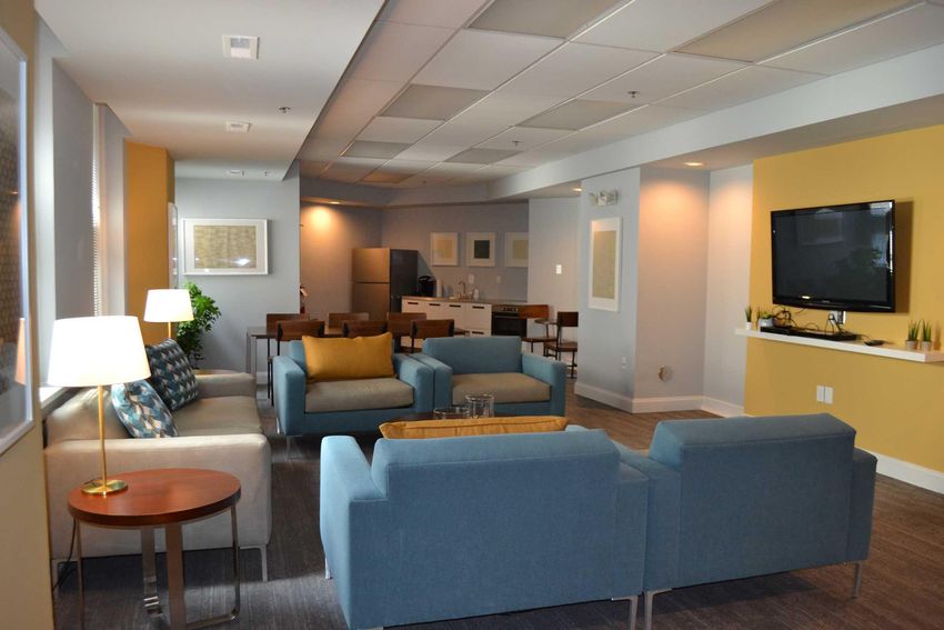 A cozy lounge area featuring modern furniture with blue and beige upholstery, a coffee table, and a floor lamp. The space includes a wall-mounted TV and a kitchenette in the background, creating an inviting atmosphere for relaxation or socializing.