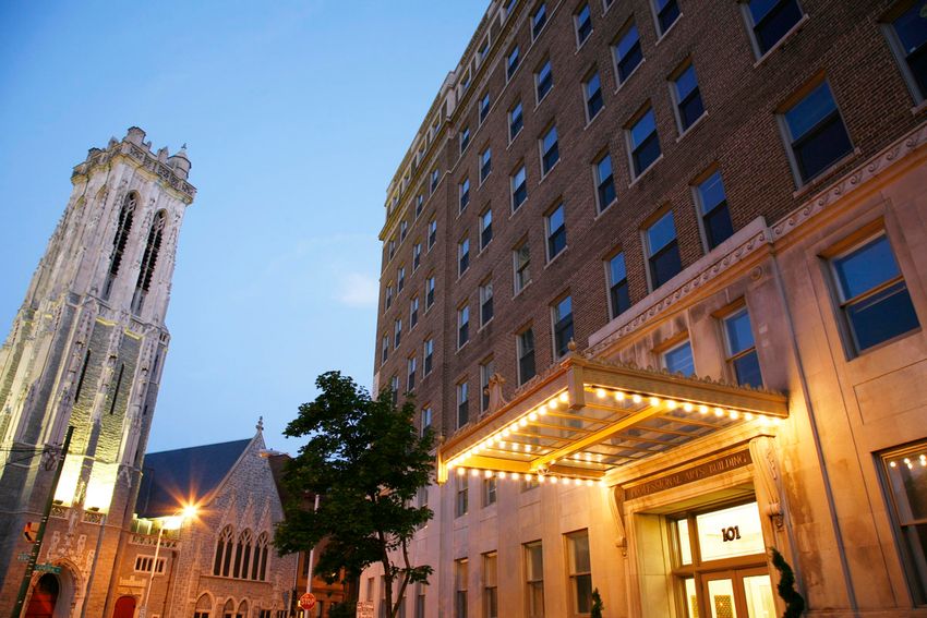 A historic building with elegant architecture, featuring a prominent entrance with lights and a marquee. To the left, a tall bell tower stands alongside a smaller church, surrounded by trees and street lights, under a twilight sky.