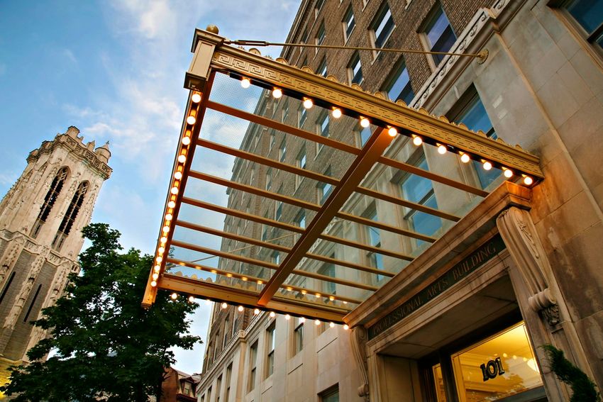 A view of the entrance to the International Arts Building, featuring a modern glass canopy adorned with lights. In the background, a tall clock tower and greenery are visible, set against a historic brick facade. The scene captures a blend of contemporary and traditional architecture.