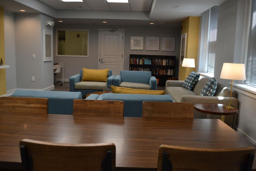A cozy lounge area featuring blue and gray sofas, a coffee table, and a wooden dining table with chairs. A lamp and bookshelves filled with books add to the inviting atmosphere, along with large windows allowing natural light into the space.