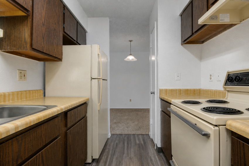 A small kitchen with wooden cabinets, a beige countertop, and appliances including a refrigerator, stove, and sink. The wall finishes are white, and there is a light fixture hanging from the ceiling. The space leads into another room with carpeted flooring visible in the background.