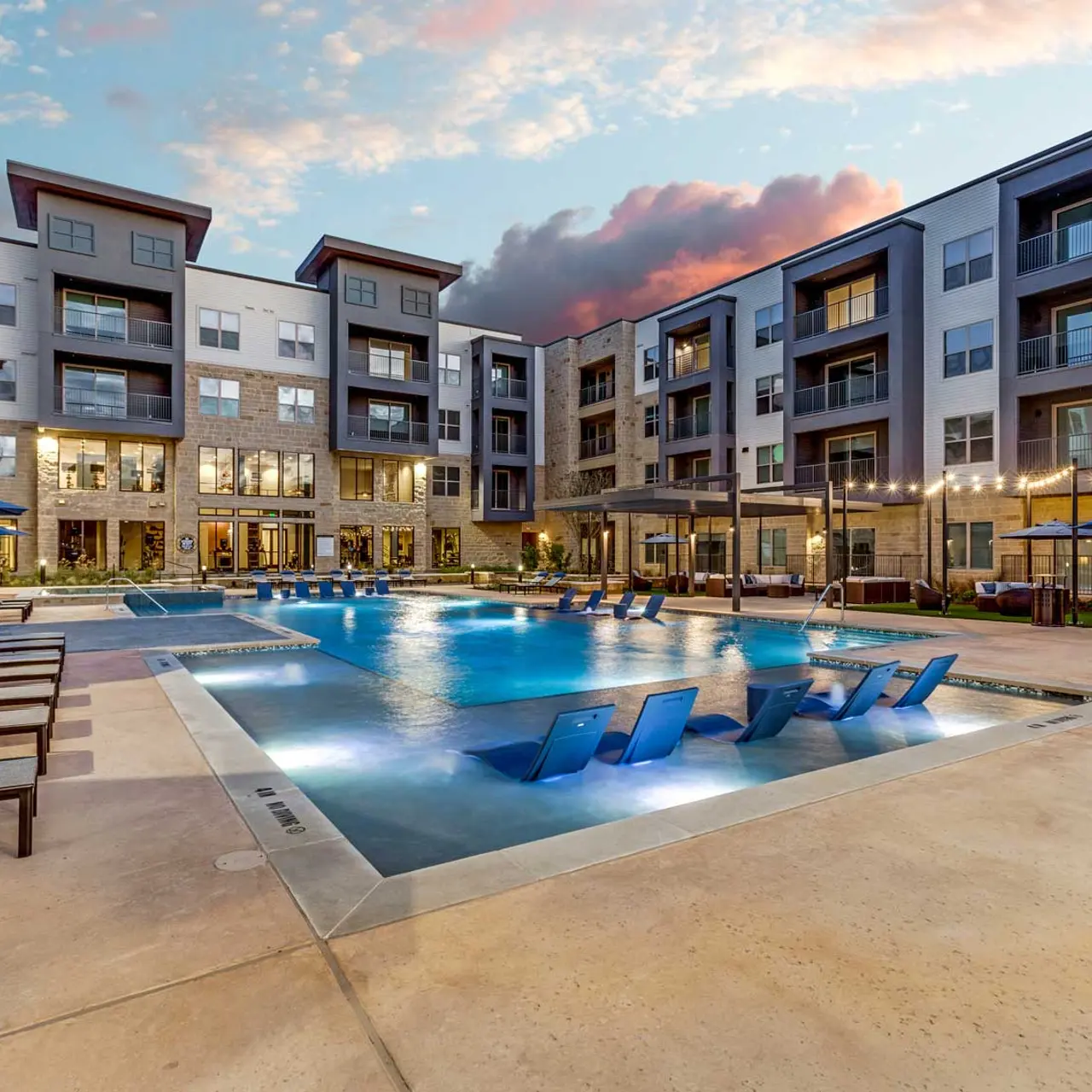 Outdoor swimming pool area at a modern apartment complex with lounge chairs and umbrellas during sunset.