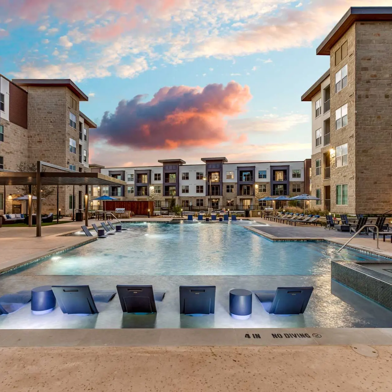 A modern apartment complex pool area featuring a large swimming pool with loungers, surrounded by buildings at sunset.