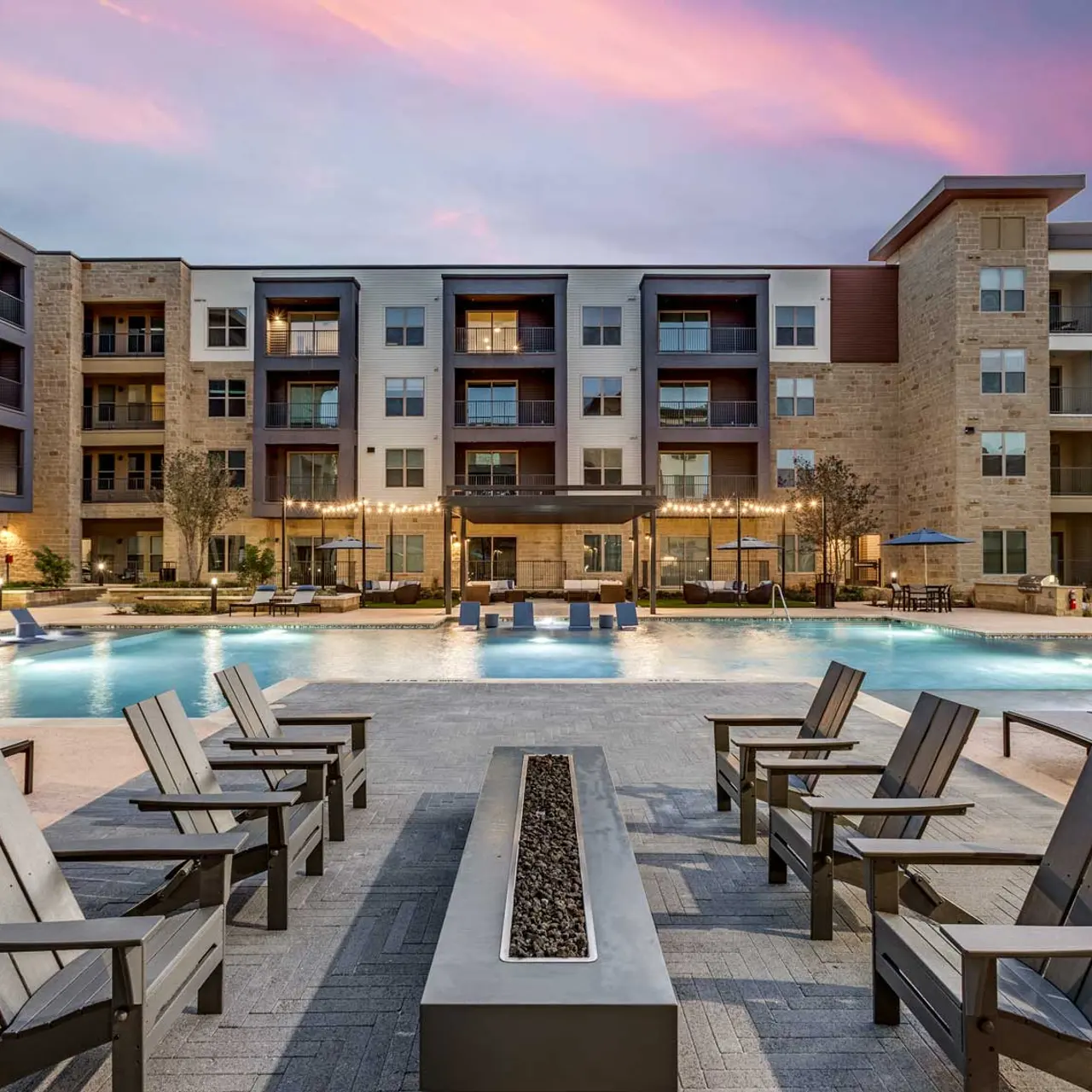 A modern apartment complex featuring a large swimming pool surrounded by lounge chairs and stylish outdoor furniture. The building has multiple floors and several balconies. The sky is painted with pastel colors during sunset.