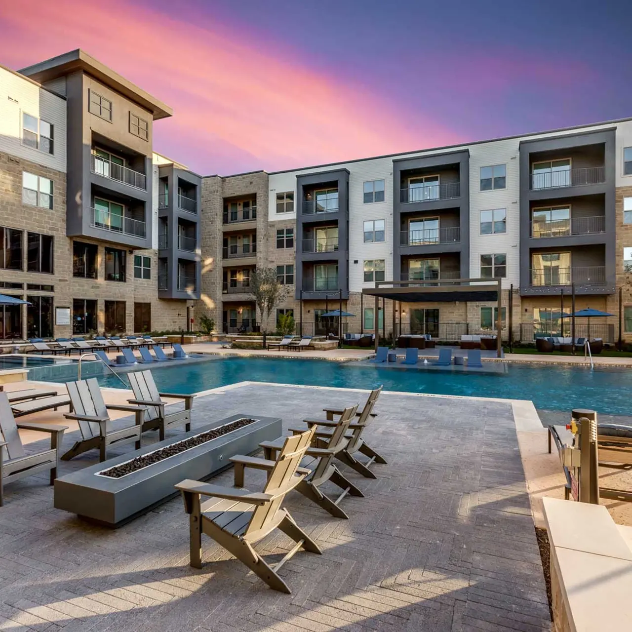 A tranquil pool area surrounded by modern apartment buildings during sunset, featuring lounge chairs and umbrellas.