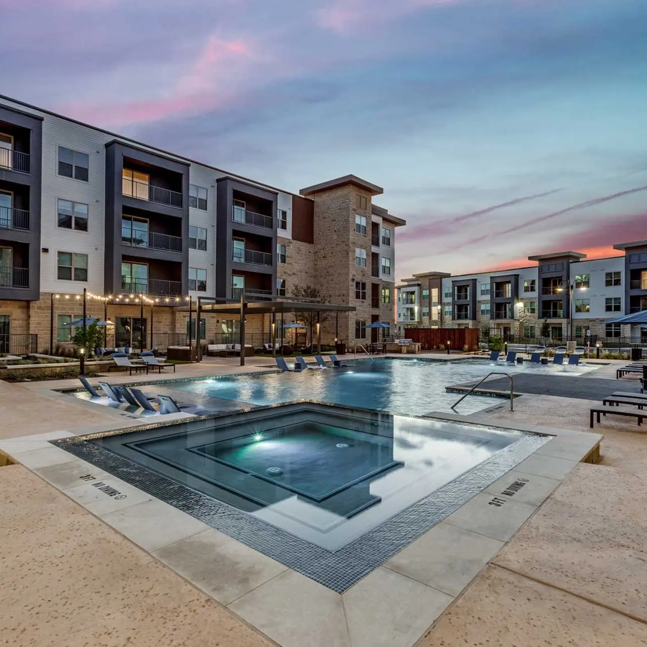 A luxurious apartment complex featuring a large swimming pool surrounded by lounge chairs and beautiful landscaping during sunset.