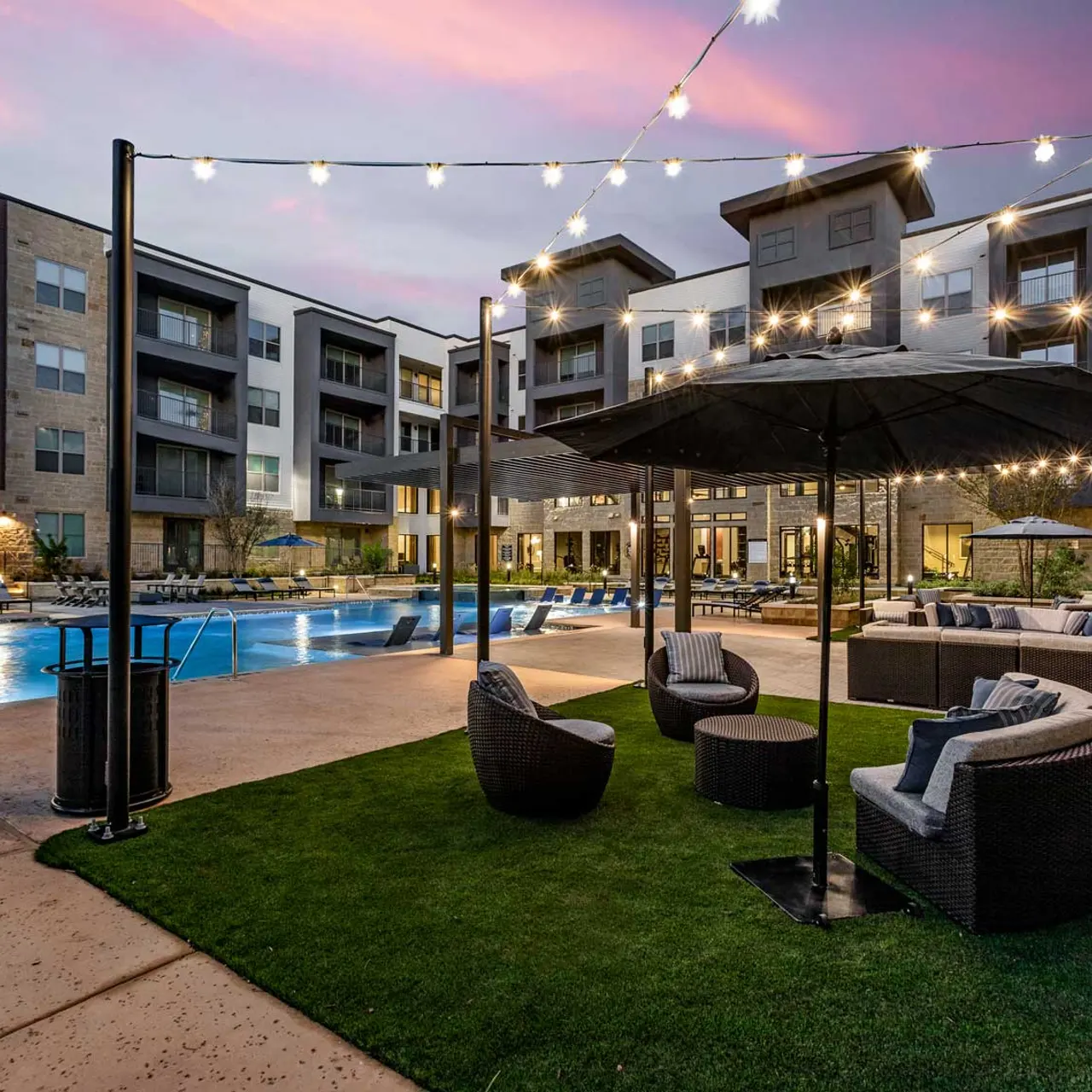 A beautifully lit apartment pool area at dusk, featuring a swimming pool, modern seating, and light strings overhead.