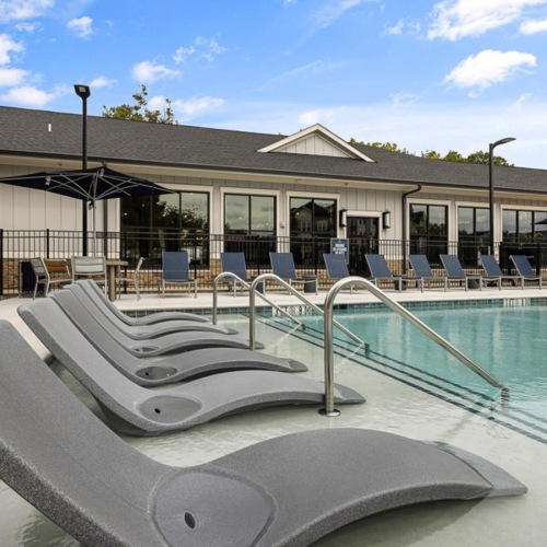 Three-Bedroom Apartments in Raleigh, NC - Conclave Glenwood - Swimming Pool with In-Water Loungers, Sun Loungers, Umbrellas, Metal Fence, Lamp Posts, and View of the Clubhouse