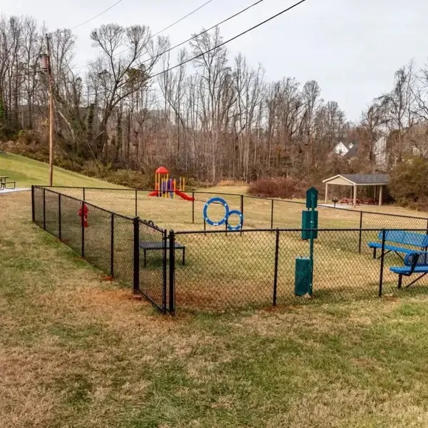 A fenced-in playground area with green grass, a small red playground structure, and blue benches. In the background, there is a swimming pool and a gazebo.