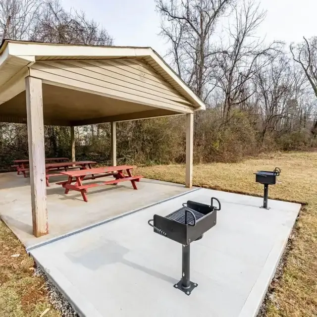 A covered picnic area with red picnic tables and built-in grills in a grassy park setting.