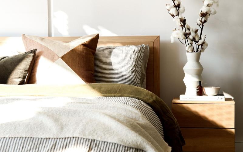 swath of sunlight shines across a well made bed and bedside table with a vase of cotton plants