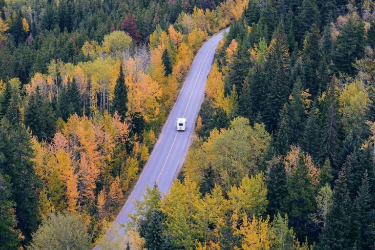 A white RV drives along a winding road through a forest with autumn-colored trees, viewed from above.