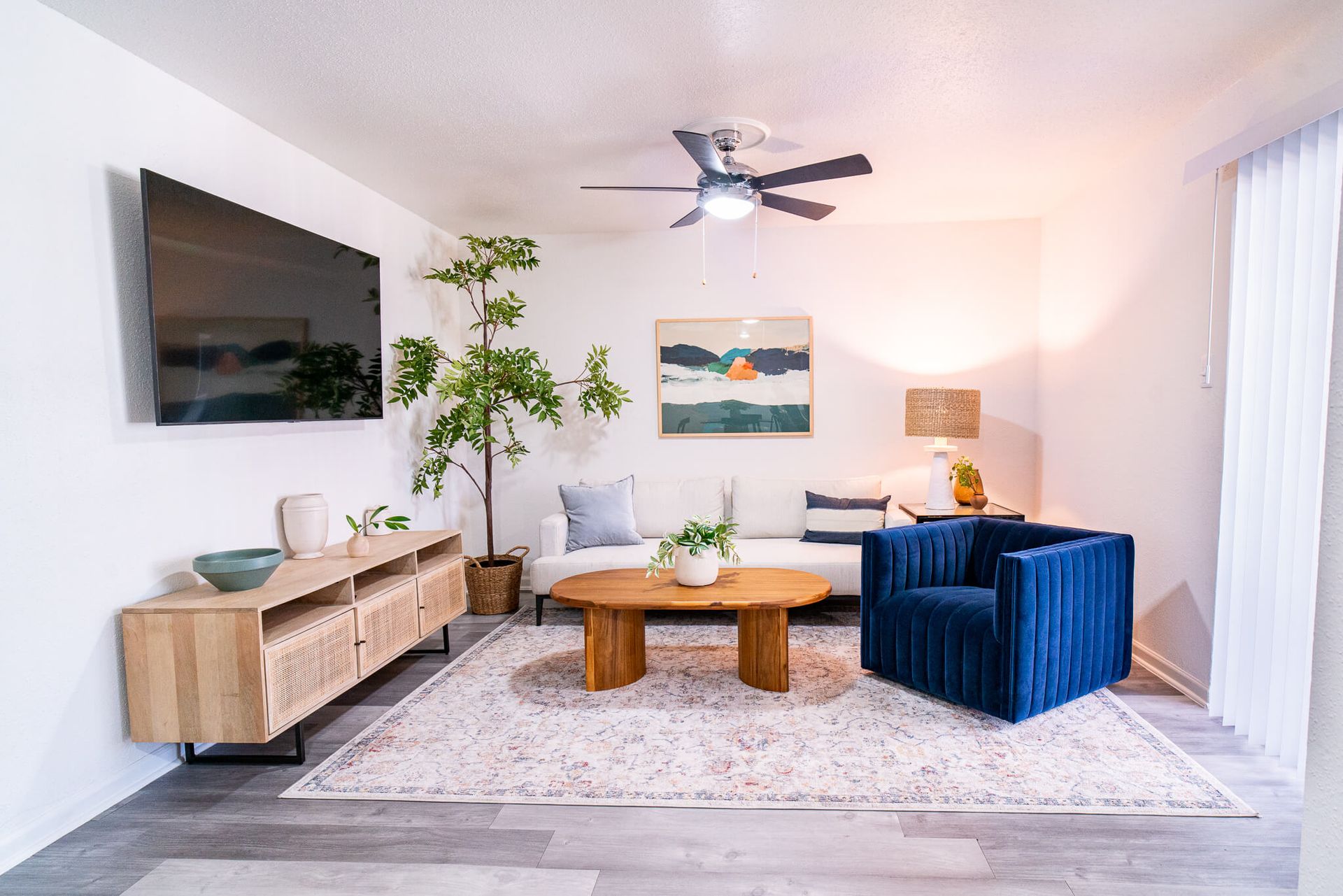 Living room in an apartment with a white sofa, blue velvet chair, wall-mounted TV, and round wooden coffee table.
