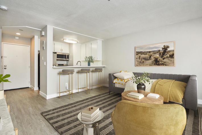 A stylish, modern living room featuring a gray sofa with yellow throw blankets, a round side table with books, and a small dining area with two bar stools at a kitchen island. The space has light-colored walls, a textured rug, and a framed artwork of a desert landscape. Bright natural light streams in through the entrance.
