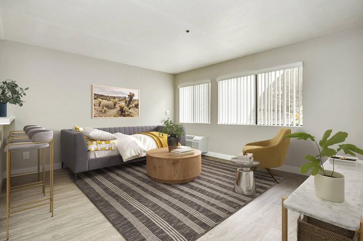 A modern living room featuring a grey sofa adorned with yellow and white pillows, a round wooden coffee table, and a yellow accent chair. Large windows allow natural light to illuminate the space, which is decorated with a potted plant and artwork on the wall. The floor is covered with a striped rug.