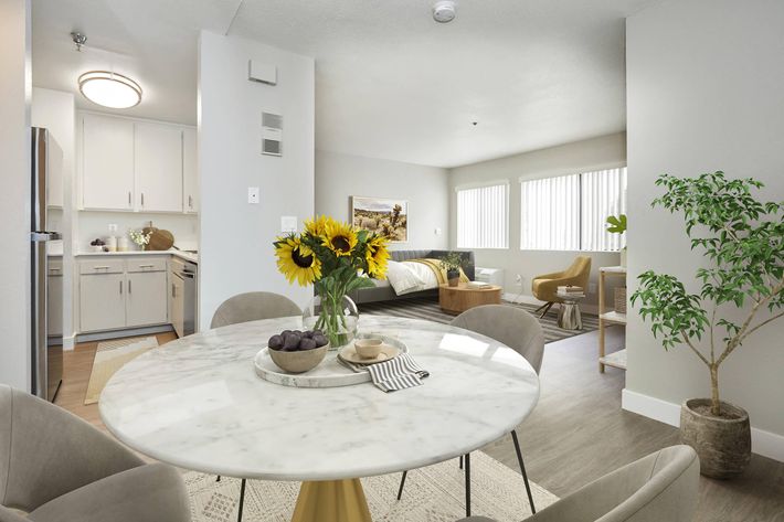 A modern, well-lit living space featuring a round marble dining table with a vase of sunflowers, a bowl of fruit, and snacks. The kitchen area is equipped with stainless steel appliances. In the background, a cozy bedroom is visible with large windows, soft furnishings, and a plant adding a touch of greenery.