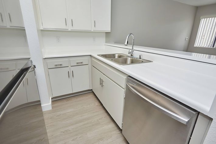 A modern kitchen featuring white cabinetry, a double sink with a sleek faucet, and stainless steel appliances. The countertop is bright and seamless, and the flooring is light wood. Natural light comes in through a nearby window, creating a clean and inviting atmosphere.