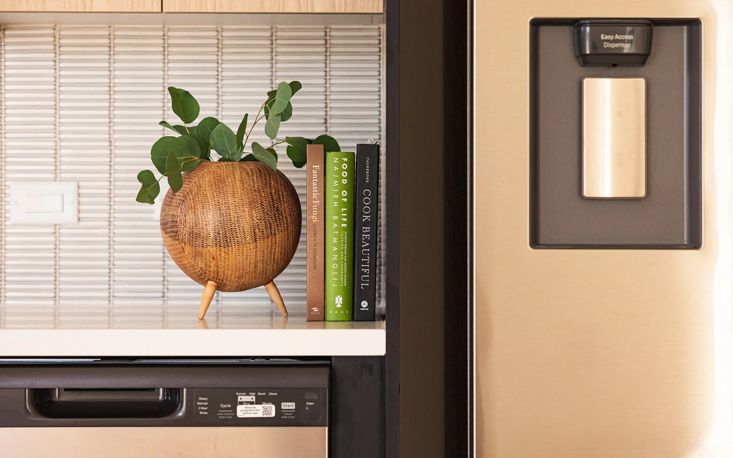 Wooden round planter with green leaves on a kitchen counter next to three cookbooks and a stainless steel refrigerator with a water dispenser.