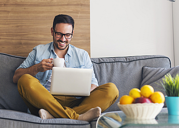 Modern living room with a joyful man sipping coffee on a stylish gray couch, accompanied by a vibrant fruit bowl and a lush green plant.