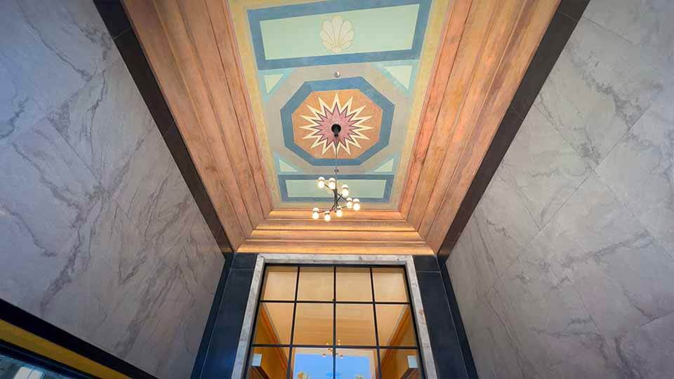 An intricately designed ceiling with geometric patterns and earthy tones, featuring a central starburst design. Below the ceiling is an ornate chandelier with spherical lights, hanging over a doorway with a glass panel, reflecting light from an unseen source.