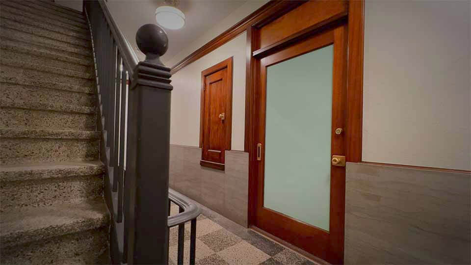 A dimly lit hallway features a carpeted staircase on the left, leading to a landing with a black metal railing. The right wall has two wooden doors, one with frosted glass and a brass handle, and the other solid wood with a brass peephole.