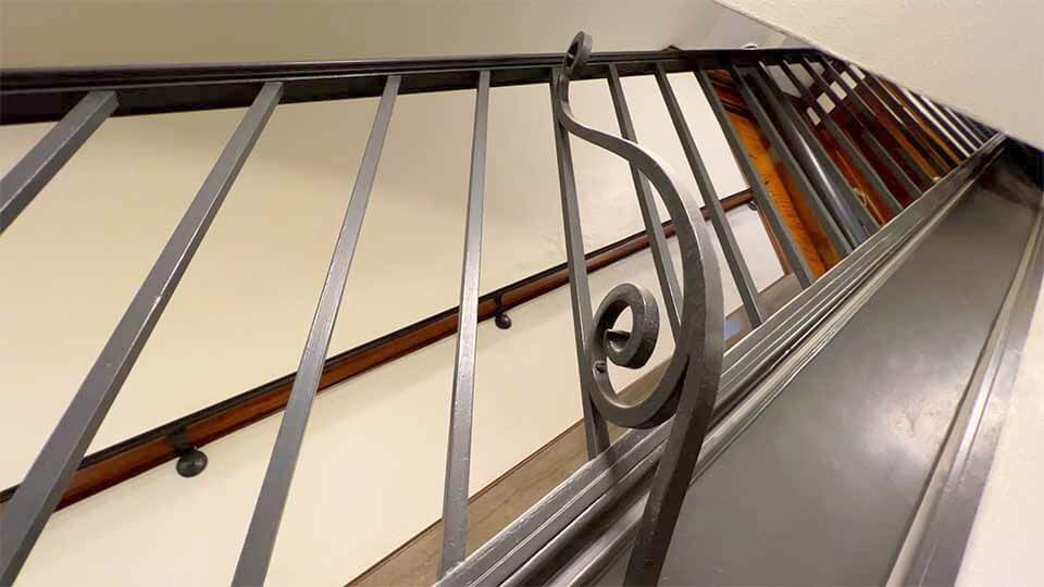 A black metal staircase railing features a decorative musical note design. The staircase walls are white, with wooden handrails running parallel to the metal railing. The image is taken from a low angle, looking up the staircase.