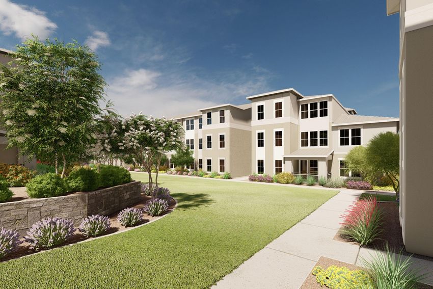 A modern residential complex featuring multiple buildings with large windows, surrounded by landscaped gardens. The pathway leads through lush green lawns with flowering plants and shrubs, under a clear blue sky with some clouds.