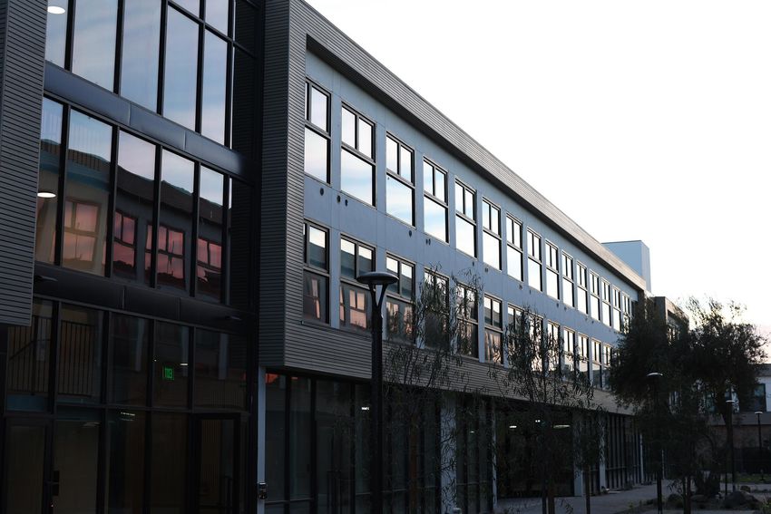 Modern building with a sleek façade featuring large windows reflecting the surrounding environment. The structure has multiple stories and is surrounded by landscaping, including small trees and lighting fixtures, set against a sunset sky.